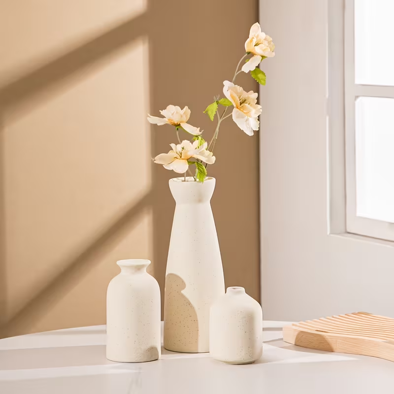 Set of three white vases with flowers on a light surface against a beige wall.