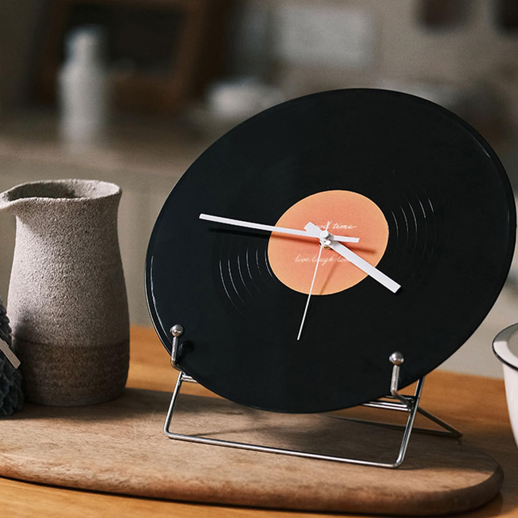 Black vinyl record clock on a stand with a brown center, placed on a wooden surface.