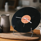 Black vinyl record clock on a stand with a brown center, placed on a wooden surface.