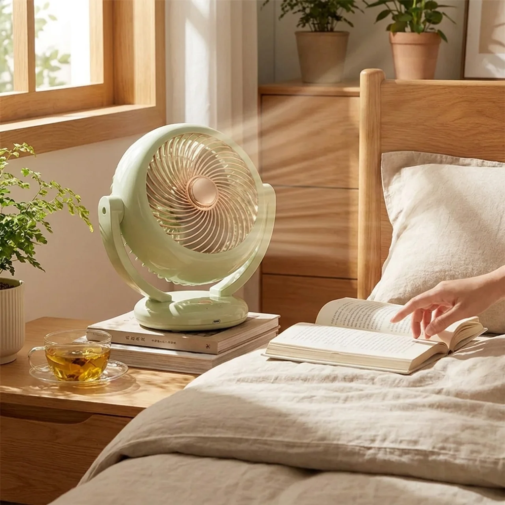 Light green fan on a wooden nightstand next to a person reading a book in a bedroom.