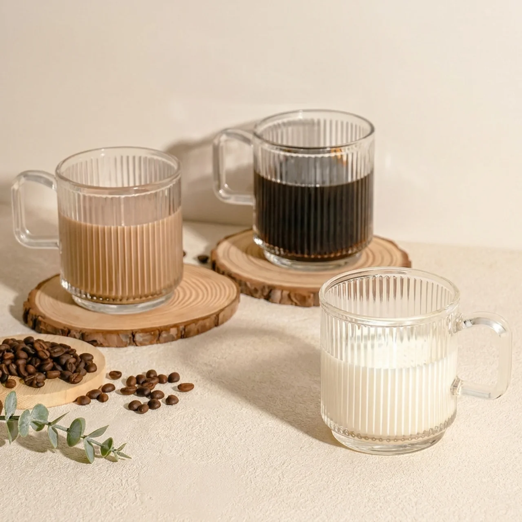Three glass mugs with different beverages on wooden coasters, surrounded by coffee beans and a sprig of eucalyptus.
