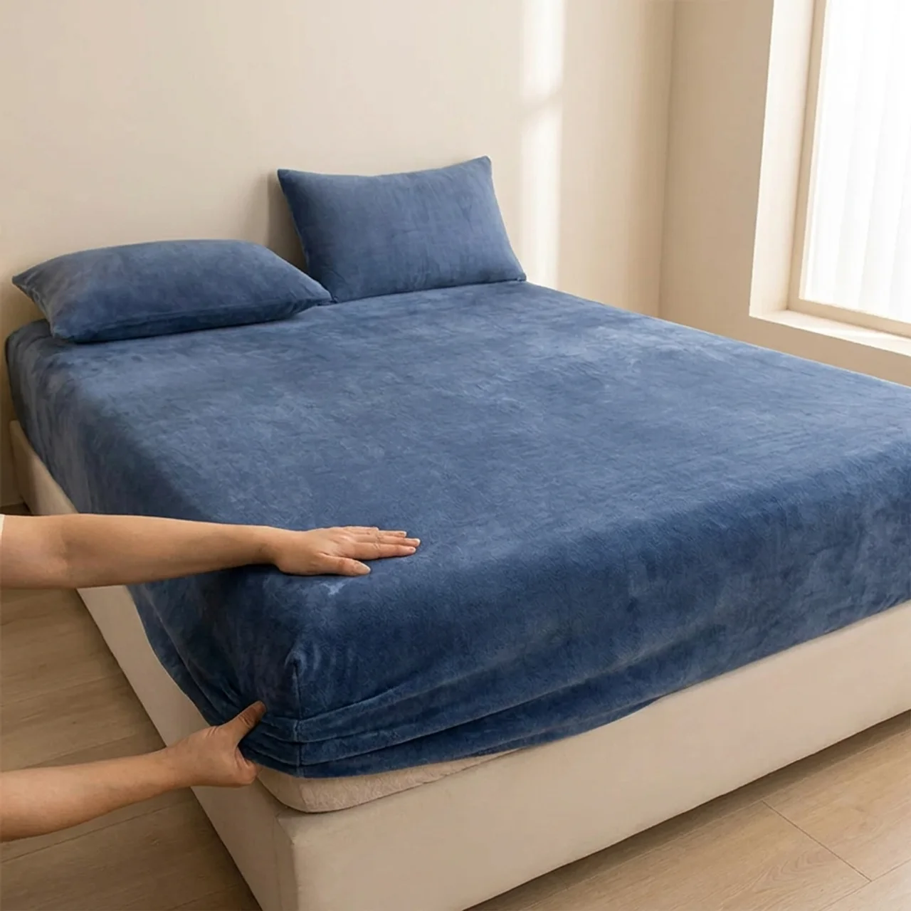 Person adjusting a blue bedspread on a bed in a room with a window.