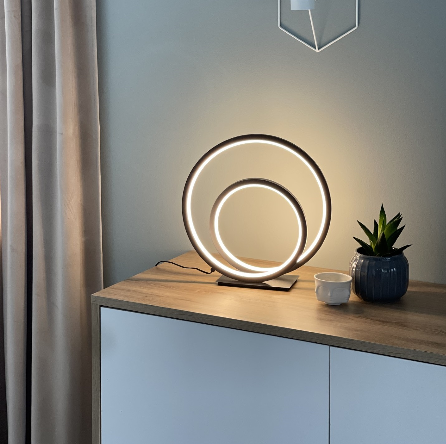 Modern table lamp with circular design on a wooden surface next to a plant and a cup.