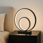Modern table lamp with circular design on a nightstand next to a bed.