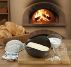 Bread-making setup with bread in a basket, dough in a bowl, and ingredients on a wooden table with a fireplace in the background.