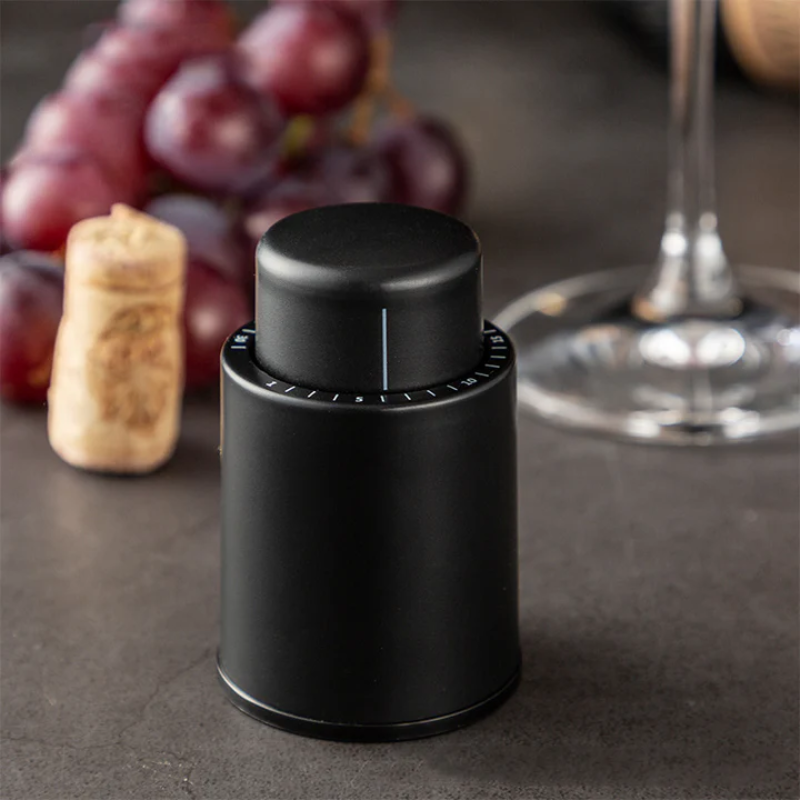 Black wine bottle stopper on a dark surface with grapes and a glass in the background