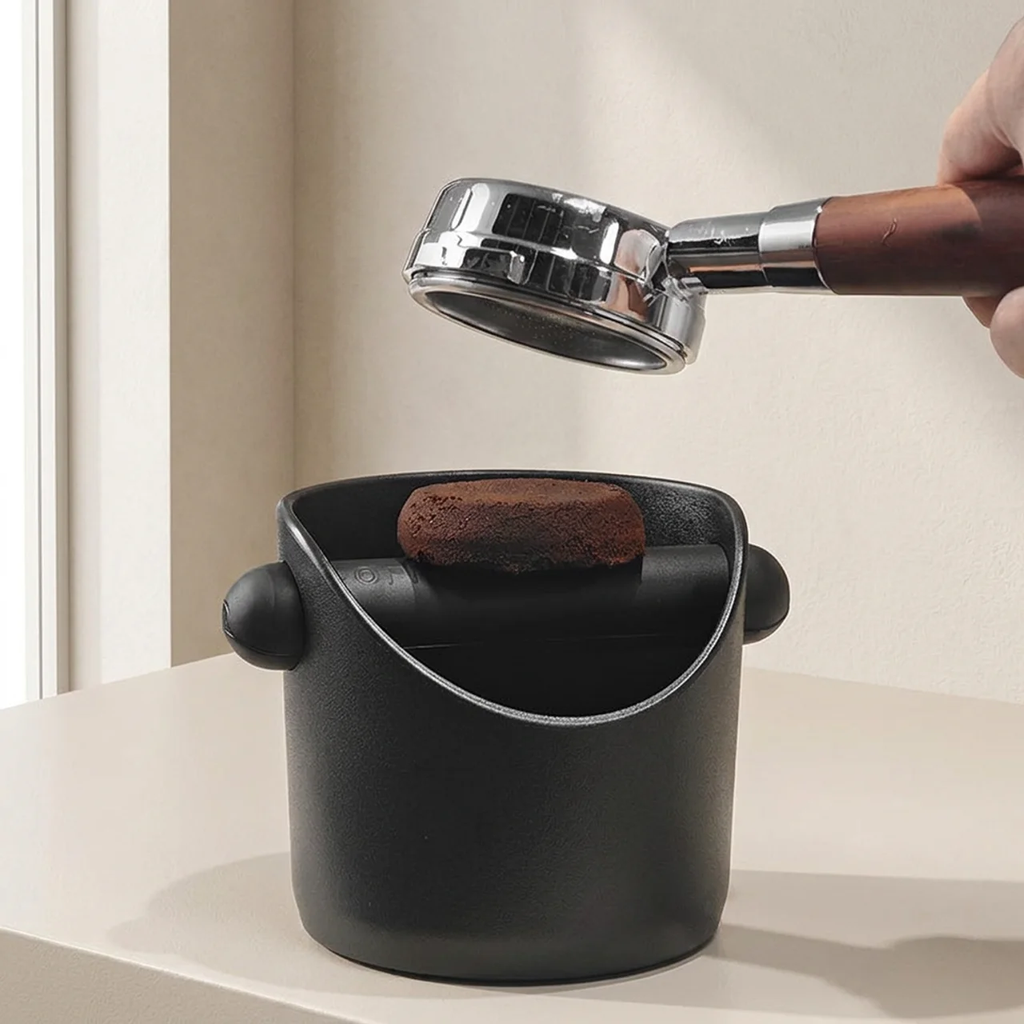 Hand holding a coffee scoop over a black container with coffee grounds on a neutral background