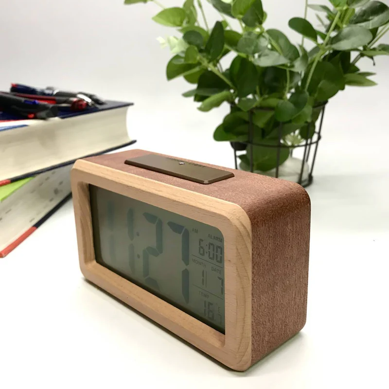 Digital clock with wooden frame on a white surface with books and a plant in the background