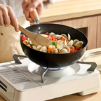 Person cooking clams and vegetables in a black wok on a portable stove.