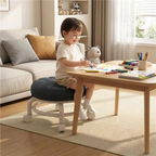 Child sitting on a black and white stool at a wooden table with art supplies in a living room.
