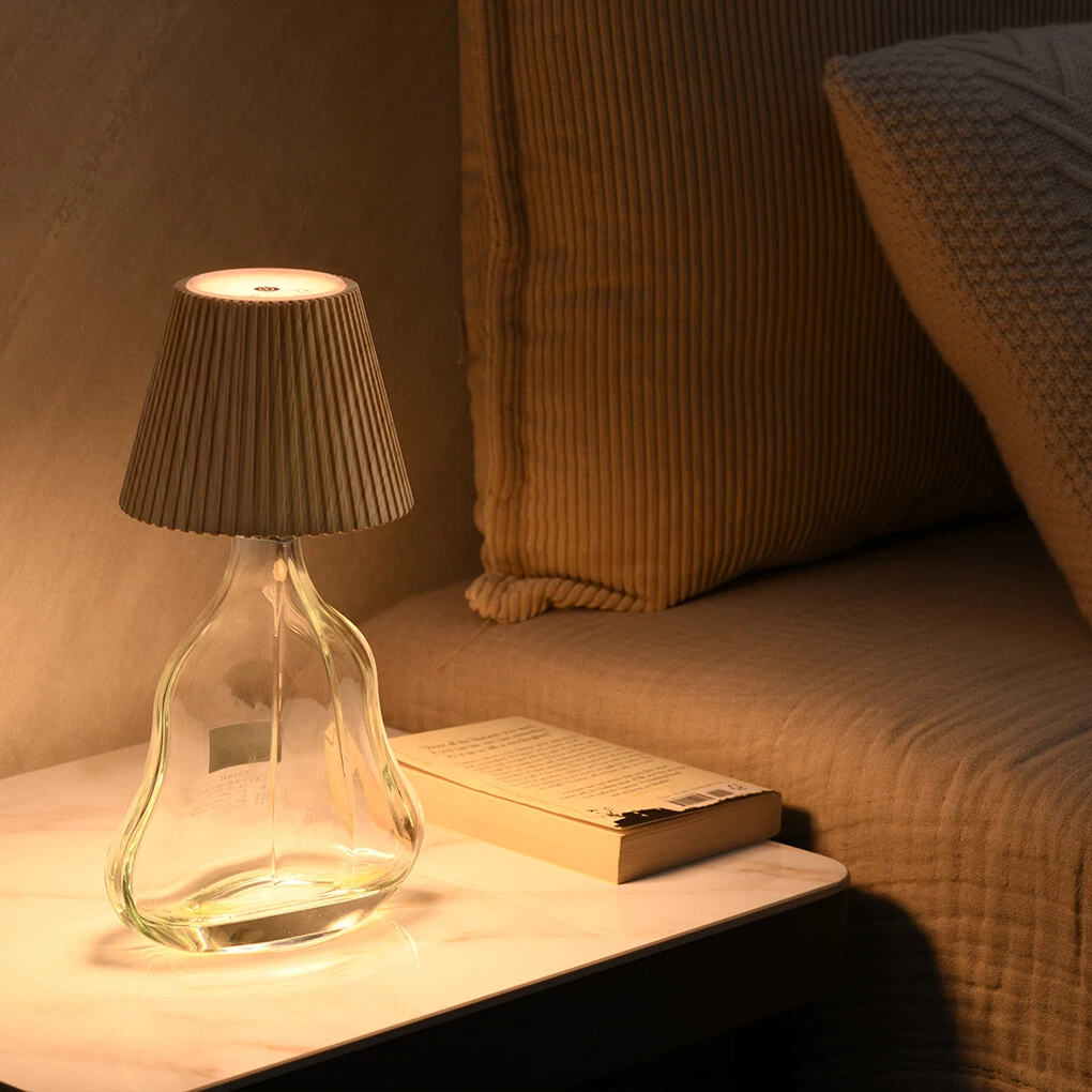 Glass table lamp with pleated shade on a marble surface next to a book, with a sofa in the background.
