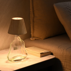 Glass table lamp with pleated shade on a marble surface next to a book, with a sofa in the background.