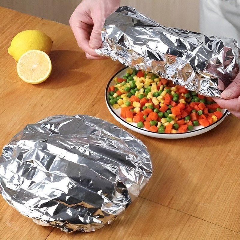 Person wrapping a plate of mixed vegetables in tinfoil on a wooden table.
