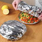 Person wrapping a plate of mixed vegetables in tinfoil on a wooden table.