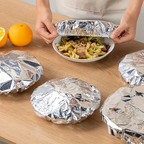 Person unwrapping a bowl of food from tinfoil on a wooden table with oranges in the background.
