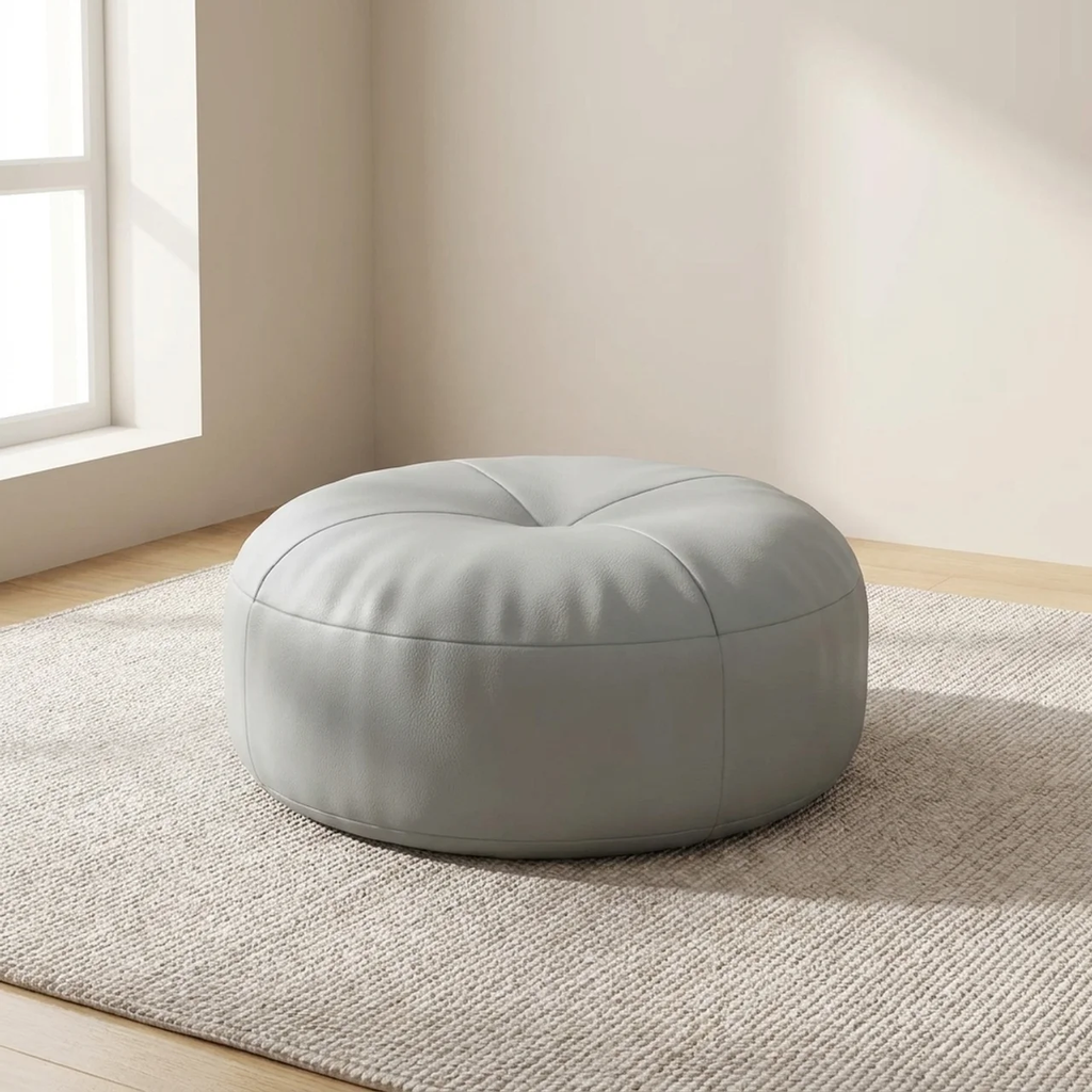 Round gray ottoman in a room with a window and beige walls.