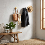 Wall with hooks holding a brown bag, black coat, and straw hat, next to a wooden bench with a plant and pillows.