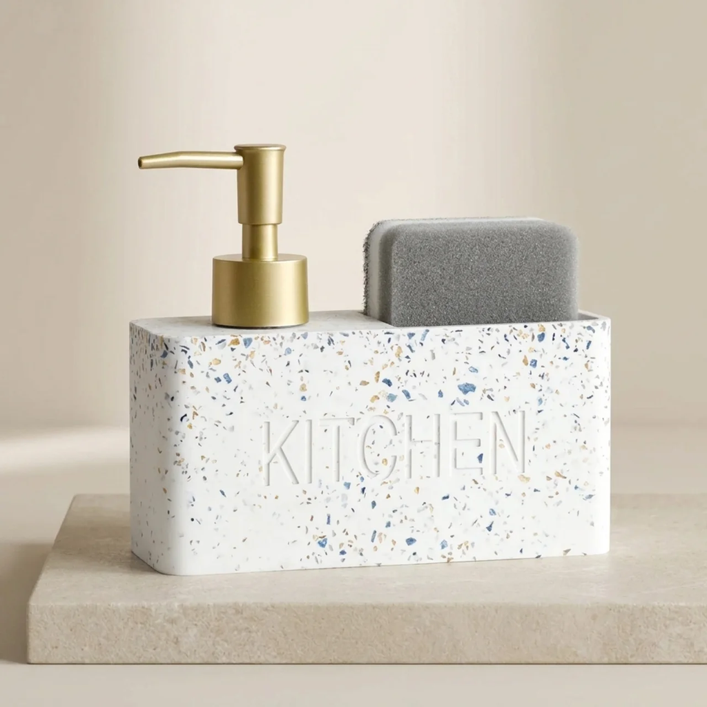 Kitchen sink caddy with gold pump, speckled container labeled 'KITCHEN', and gray sponge on a beige surface.