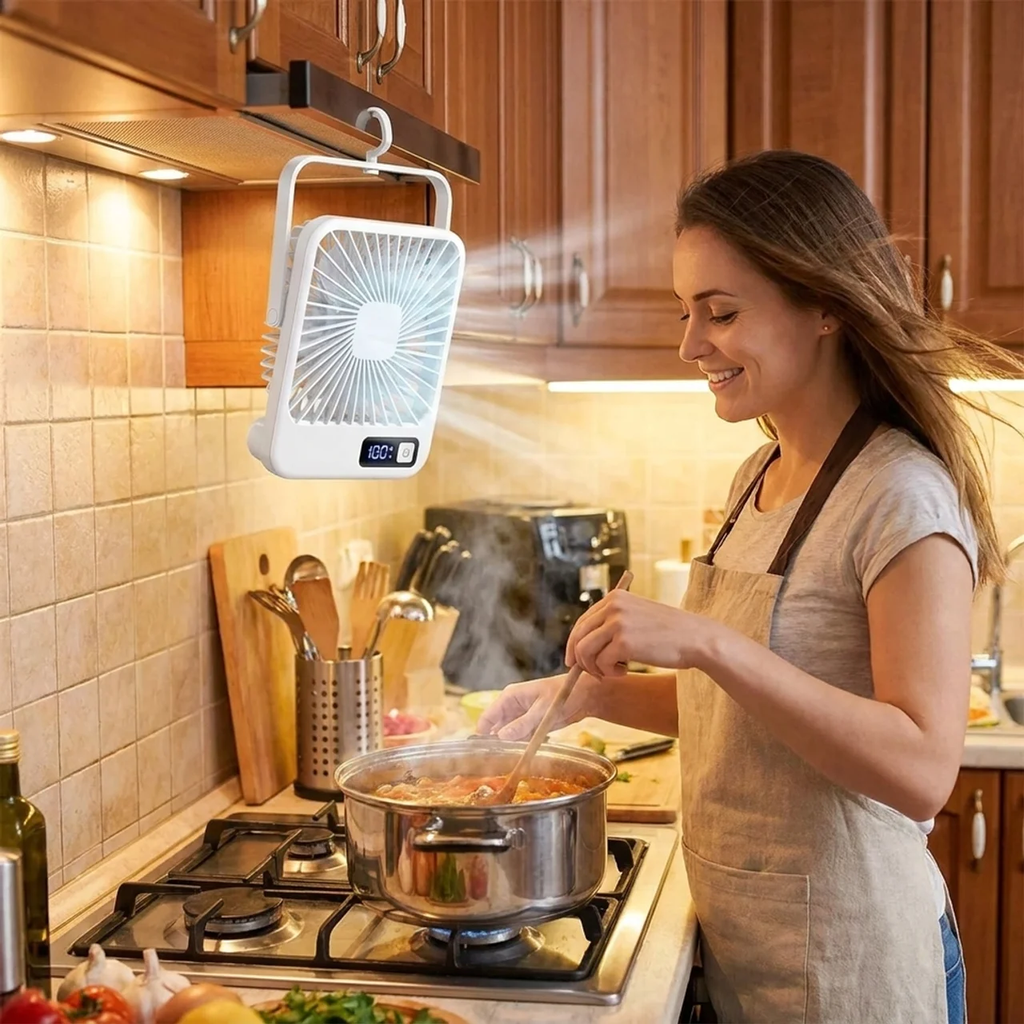 A woman in a beige apron stirs a pot on a gas stove in a warm kitchen, while a white portable fan hangs from a cabinet hook above her.