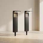 Two modern outdoor bollard lights in a matte black finish, standing on a light beige stone floor against a neutral beige wall.