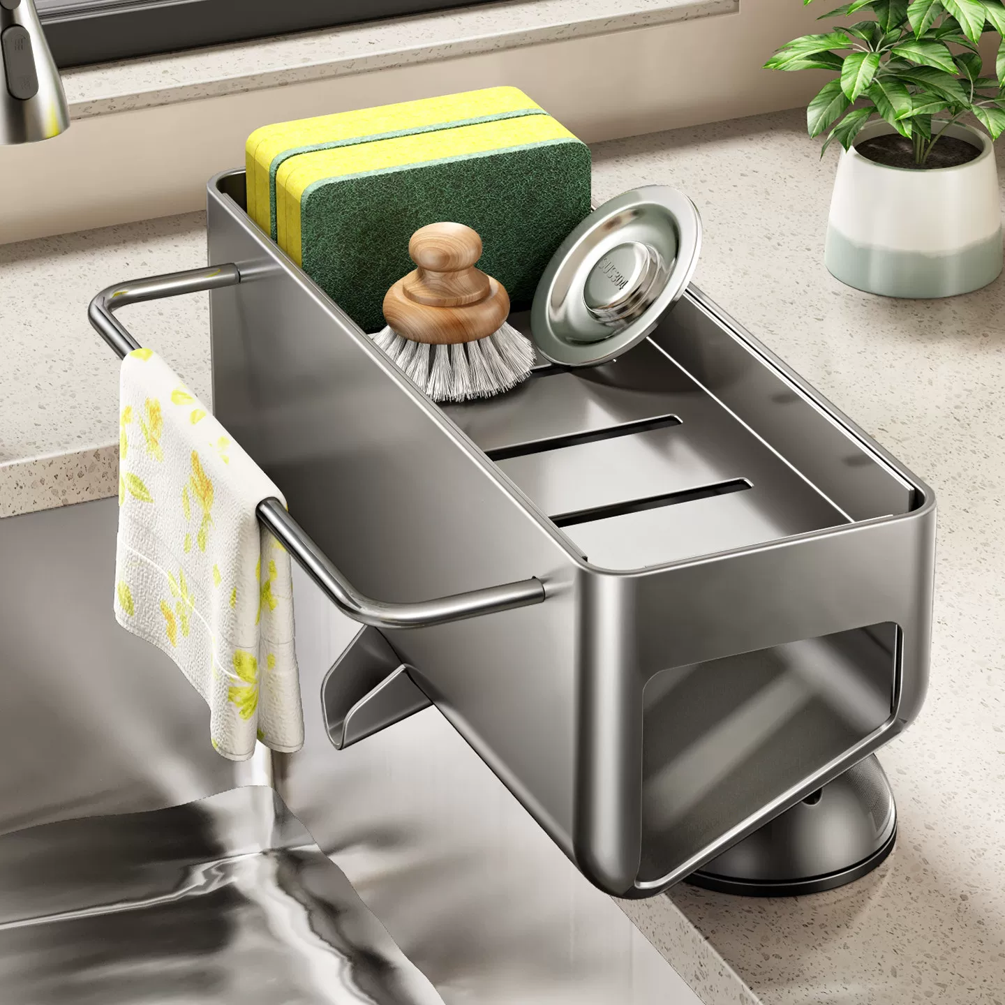 A stainless steel kitchen sink organizer with a suction base, shown mounted over a sink. The organizer holds a green and yellow sponge, a scrubber, a metal lid, and a white towel with yellow floral print.