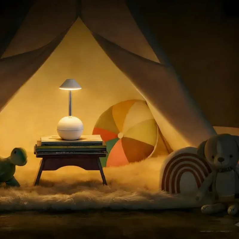 A cozy children's play scene under a canvas tent, featuring a small round white table lamp on a stack of books, a plush teddy bear, a rainbow pillow, and a colorful ball toy.