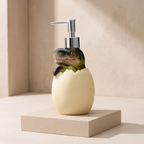 A dinosaur-themed soap dispenser featuring a realistic green and black dinosaur peeking out of a speckled cream-colored egg-shaped base, with a chrome pump. The dispenser is displayed on a matching beige pedestal against a neutral beige background.
