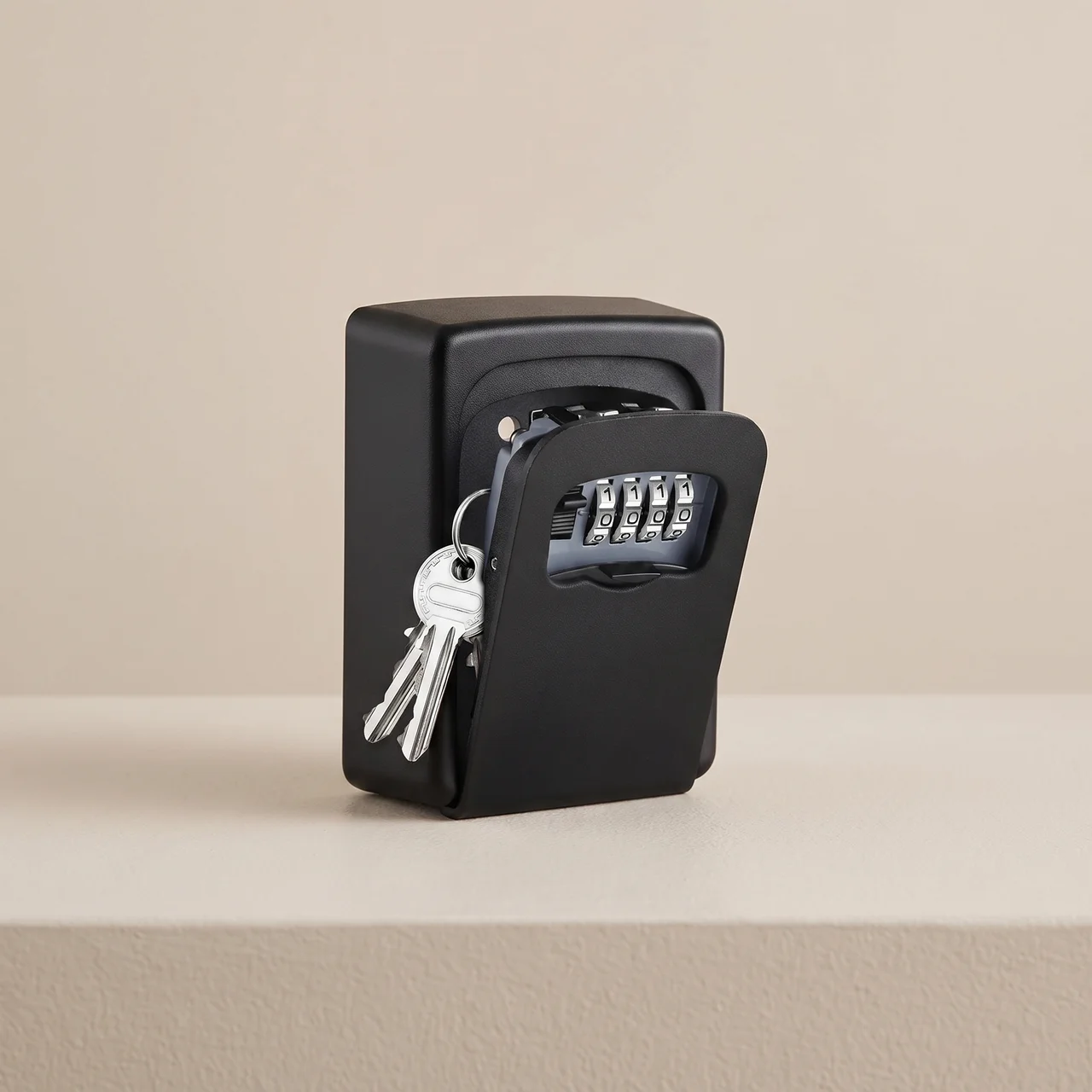 A black key safe with a four-digit combination lock and attached keys, shown open on a beige surface against a matching beige background.