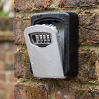 A waterproof outdoor wall-mounted key box with a sliding silver-gray lockable door and four combination dials, shown mounted on a weathered brick wall with water droplets on the surface.
