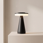 A modern table lamp with a matte black tapered base and a circular black top emitting a warm white glow, set on a light beige pedestal against a neutral beige wall.