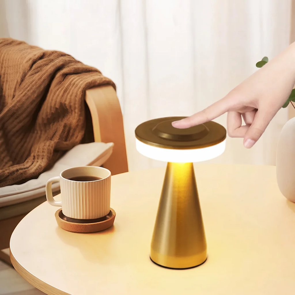 A modern table lamp with a gold metallic cone base and a circular light top, shown on a light wood table in a cozy indoor setting. A hand is touching the top of the lamp, and a cup sits nearby on a coaster.