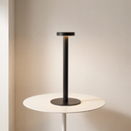 A modern black metal table lamp with a circular base and a slim cylindrical stem, shown lit on a white round side table against a warm beige wall.