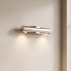 A modern cylindrical wall sconce with a brushed silver finish, mounted on a warm beige wall and casting a soft downward light.