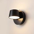 A modern matte black wall sconce light fixture with a cylindrical shade and circular wall mount, shown illuminated against a warm off-white wall.