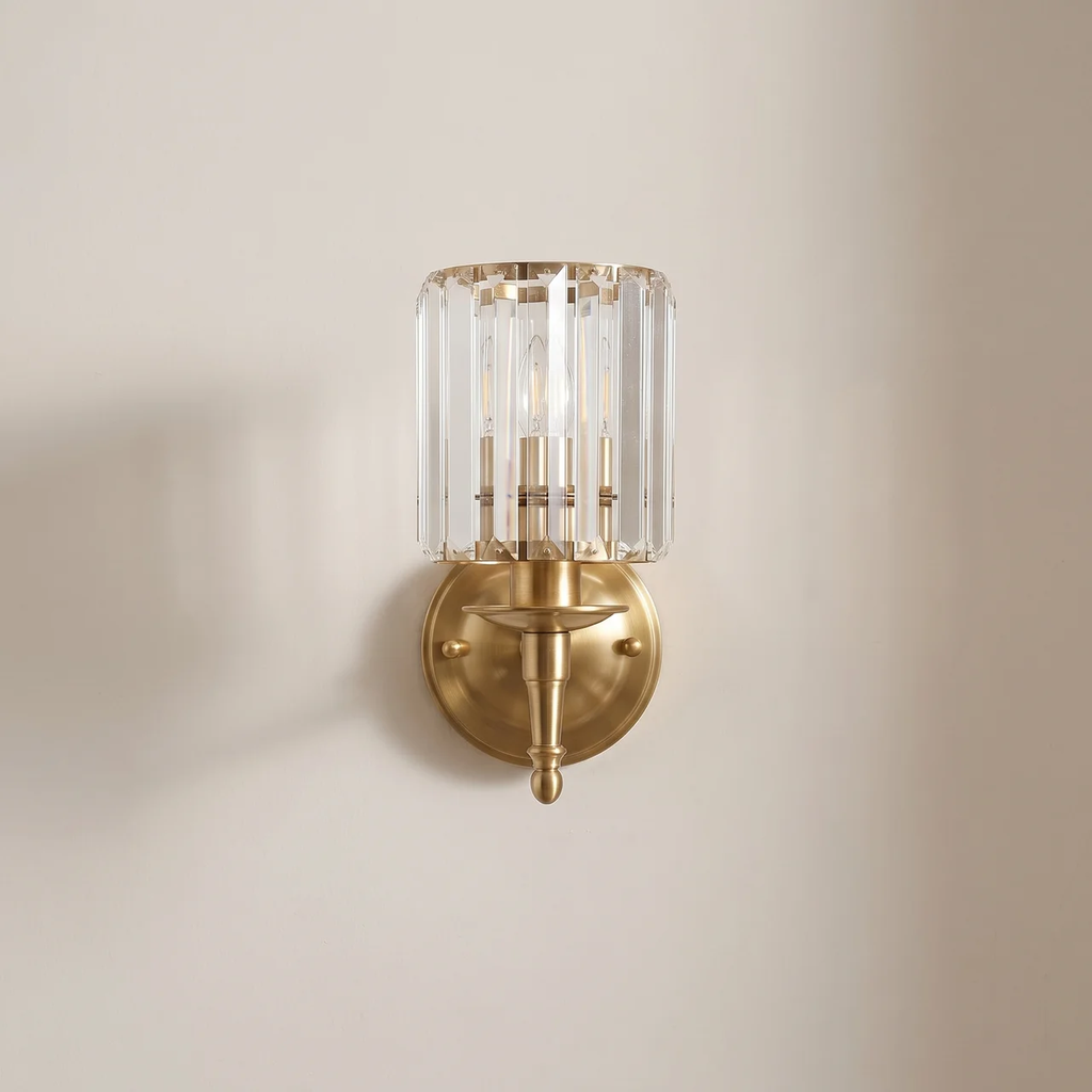 A wall-mounted sconce with a brushed gold metal base and a clear cylindrical crystal shade, shown against a plain beige wall.
