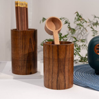 Two dark brown wooden cylindrical holders on a white surface, one holding pencils and the other holding a wooden scoop, with soft-focus greenery and a blue textured object in the background.