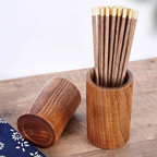 A wooden chopstick holder set on a wooden tabletop, with one upright cylinder filled with brown wooden chopsticks that have gold-toned tips, and another matching cylinder lying on its side. A small navy fabric corner with white floral print is visible at the lower left.