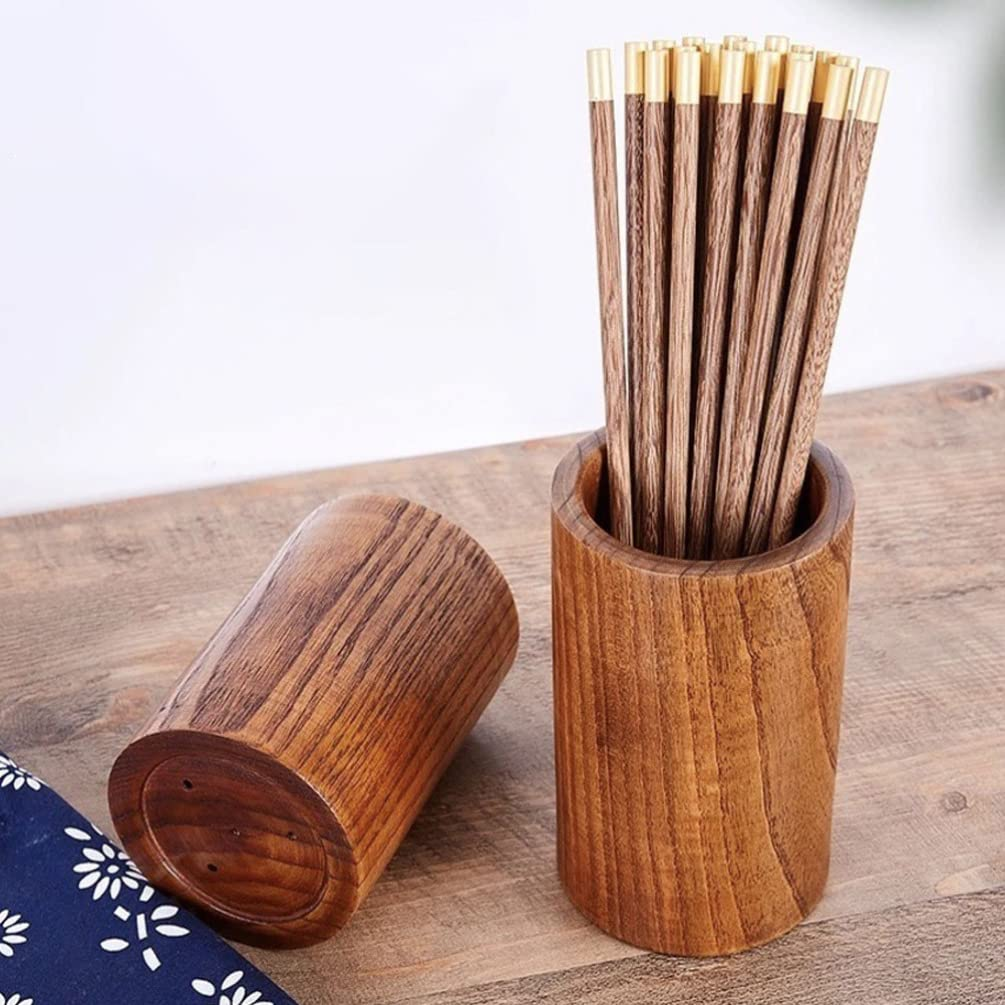 A wooden chopstick holder set on a wooden tabletop, with one upright cylinder filled with brown wooden chopsticks that have gold-toned tips, and another matching cylinder lying on its side. A small navy fabric corner with white floral print is visible at the lower left.