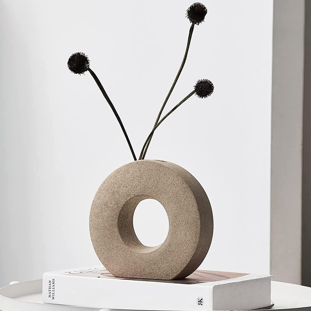 A minimalist beige donut-shaped ceramic vase with a central hole, holding three long black flower stems with round dark blooms, displayed on top of a white book against a clean light gray background.