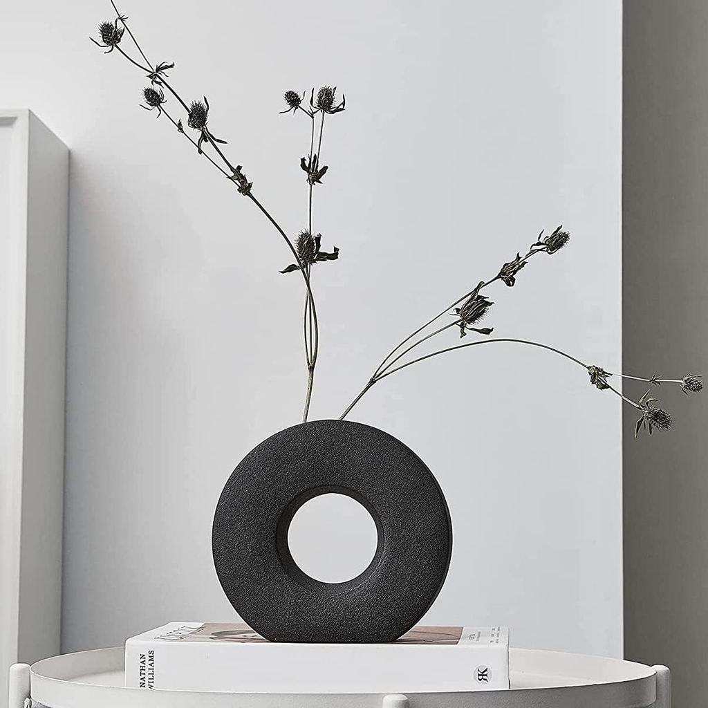 A matte black donut-shaped vase holding dried black flower stems, displayed on a white side table against a light gray wall.