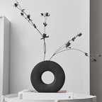 A matte black donut-shaped vase holding dried black flower stems, displayed on a white side table against a light gray wall.