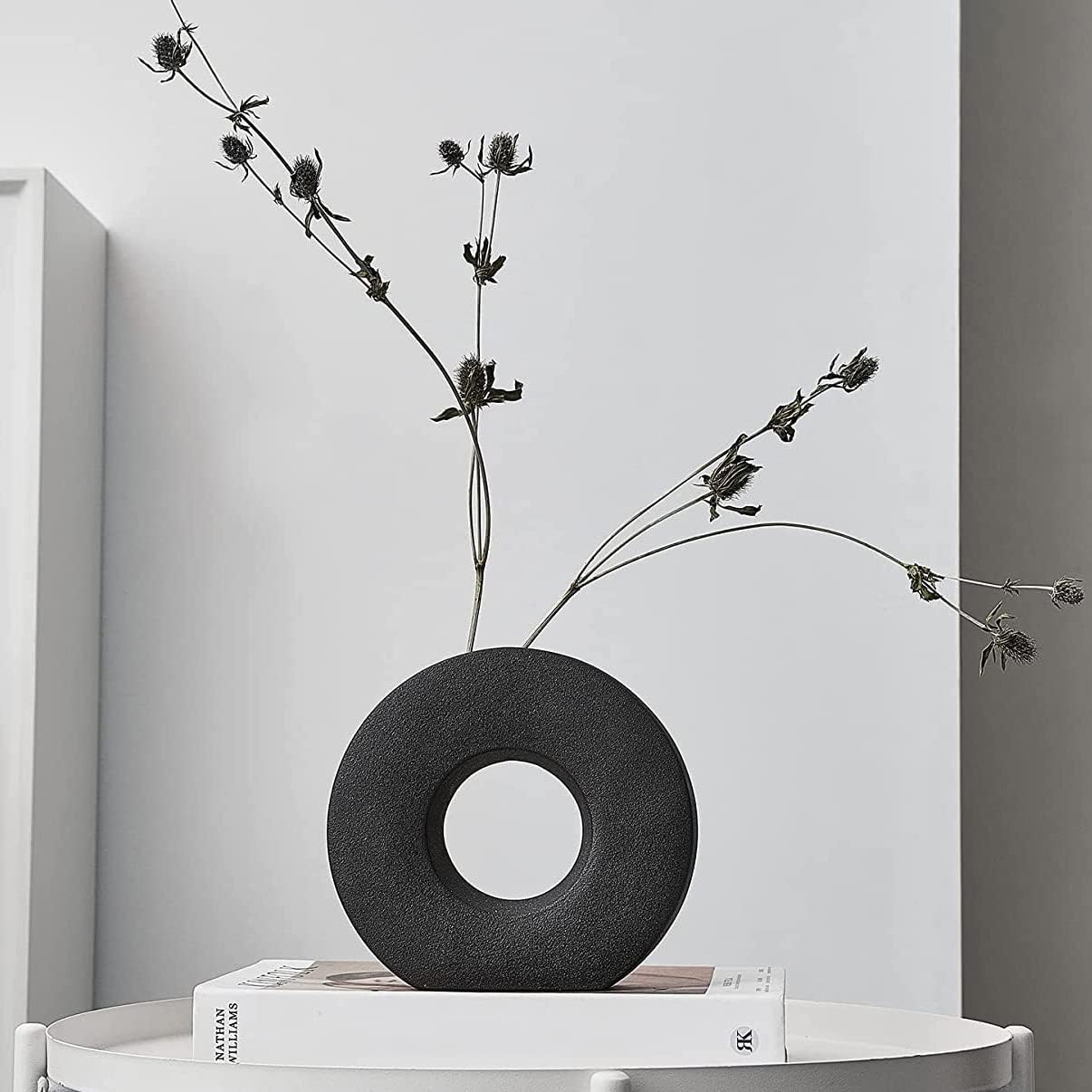 A matte black donut-shaped vase holding dried black flower stems, displayed on a white side table against a light gray wall.