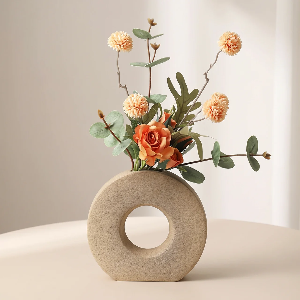 A beige donut-shaped ceramic vase with a circular cutout, holding an artificial floral arrangement with orange roses, peach pom-pom flowers, and green eucalyptus leaves, set on a light cream surface against a soft neutral background.