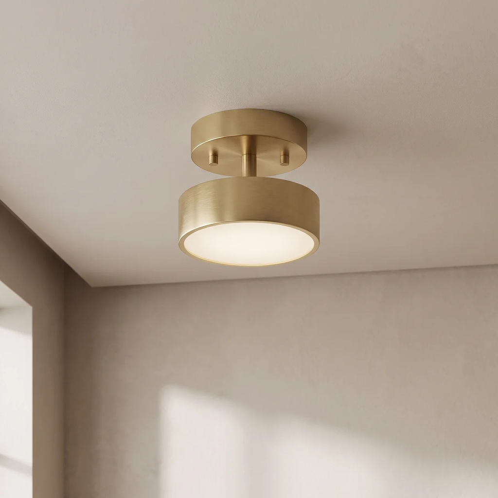A modern semi-flush mount ceiling light fixture in a brushed gold finish, emitting a warm glow from its circular shade, shown installed on a textured off-white ceiling in a minimalist room corner.