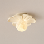 A modern ceiling light fixture with a flower-like cream shade and a glowing spherical moon-shaped bulb, shown against a plain beige wall.
