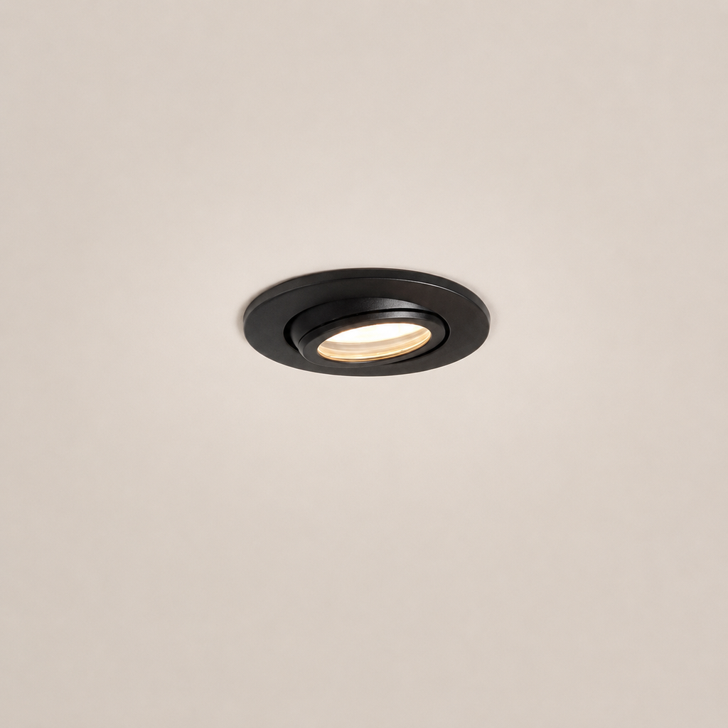 A round recessed ceiling light fixture in matte black, shown installed against a plain off-white ceiling with the light turned on, emitting a warm glow.