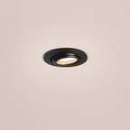 A round recessed ceiling light fixture in matte black, shown installed against a plain off-white ceiling with the light turned on, emitting a warm glow.