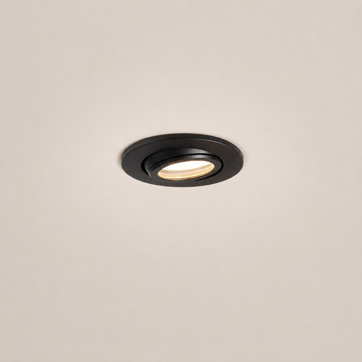A round recessed ceiling light fixture in matte black, shown installed against a plain off-white ceiling with the light turned on, emitting a warm glow.