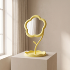 A yellow flower-shaped tabletop mirror with a sculptural stem base, displayed on a white pedestal in a softly lit room with beige walls and curtains.