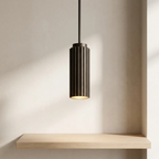 A modern cylindrical pendant light with a dark bronze fluted shade and a warm glowing light, suspended from a black cord above a light wood floating shelf against a beige wall.